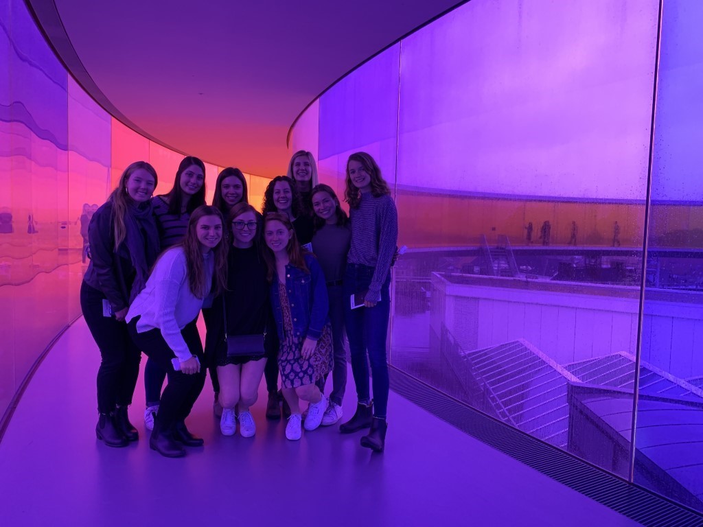 Ten young women stand in a group in a hallway which has walls of purple glass which gradually changes color to be pink, then orange in the background. They are smiling at the camera].