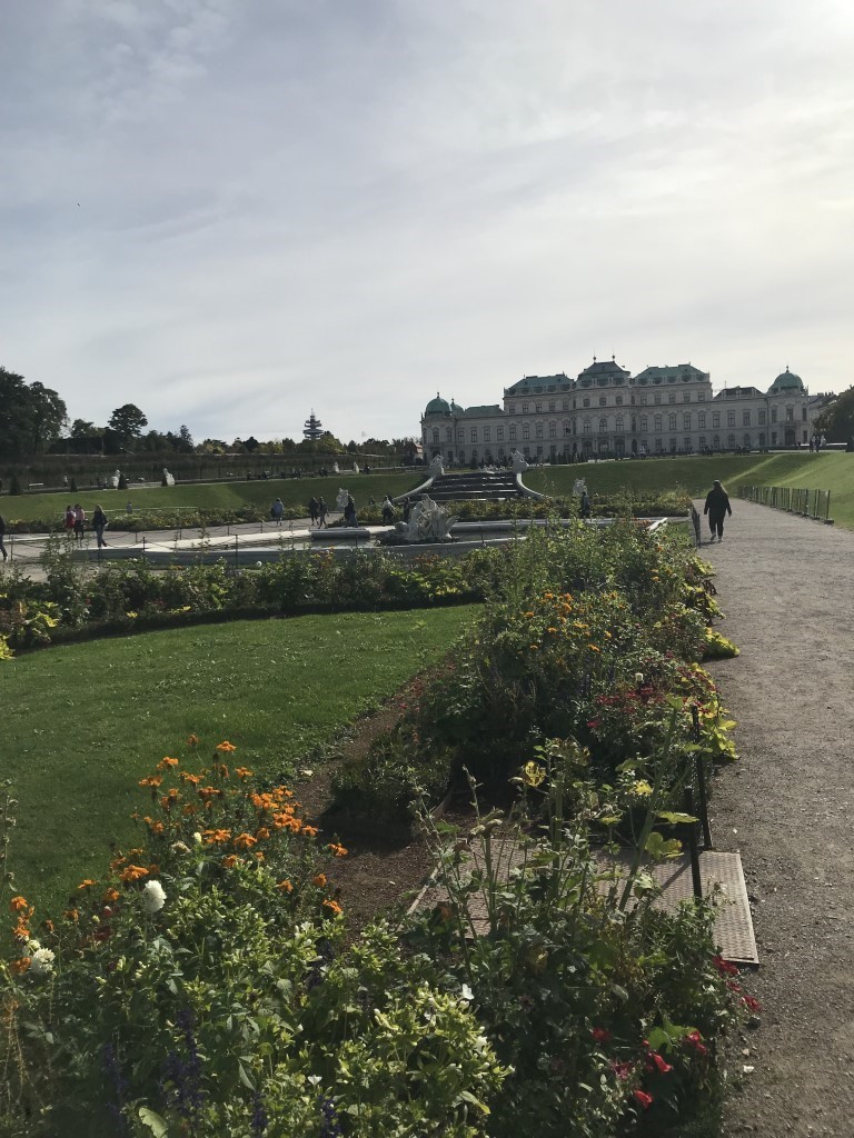 An elegantly landscaped garden of orange, red, and yellow flowers, green plants, and a fountain lead up to a dramatic staircase to a large white palace with a green roof. Several people wander about the garden on gravel paths.