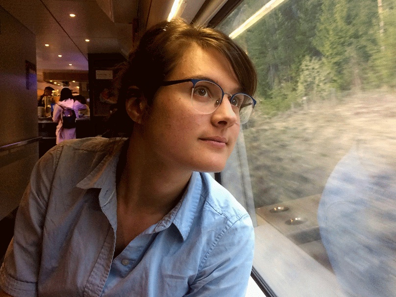A woman in a light blue shirt looks out the window of a train as wilderness rushes by.