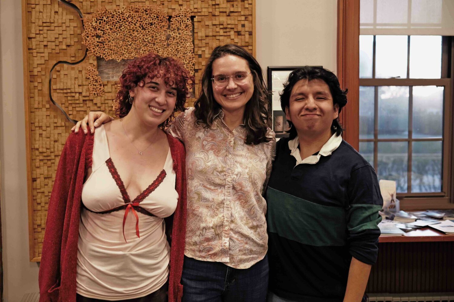 One young woman with curly hair, a woman with long brown hair, and a young man with dark hair stand with arms around each others shoulders in front of a wooden piece of art.