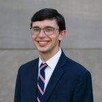 Ben Sheriff, a white man with brown hair and glasses in formal wear, smiles at the camera while standing in front of a stone background.