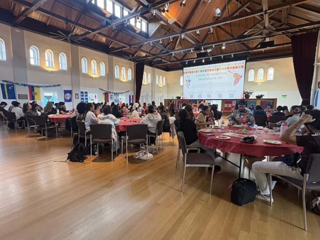 Groups seated at round tables watch a screen with the words "Latine Affirmation Month Kick-Off"