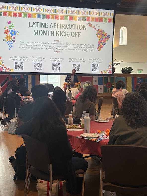 Groups seated at round tables watch a screen with the words "Latine Affirmation Month Kick-Off"